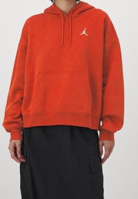 Sudadera roja de tela suave, con un bolsillo tipo canguro, puños de canalé y un logo blanco de Jordan en el pecho. Llevada sobre una falda negra.