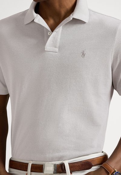 Ανοιχτό γκρι πόλο μπλουζάκι με υφασμάτινη υφή, γιακά με δύο κουμπιά και διακριτικό κεντημένο λογότυπο. Συνδυάζεται με καφέ δερμάτινη ζώνη.