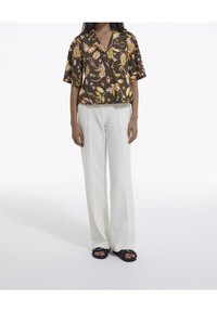 Camicia a maniche corte con bottoni e motivo floreale in nero, giallo e arancio. Abbinata a pantaloni bianchi larghi e sandali neri.
