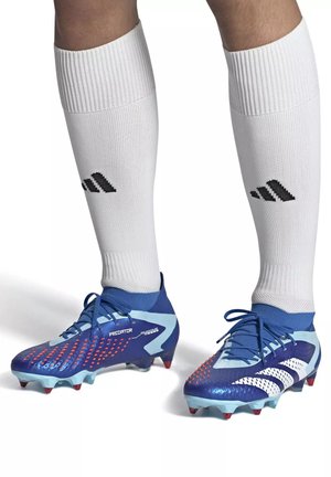 Jambes portant des chaussettes montantes blanches Adidas et des crampons de football Adidas Predator bleus avec des crampons rouges sur fond blanc.