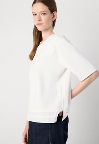 Hvit kortermet topp med rund hals, sidespalter og subtil tekstur. Kombinert med mørke denimjeans, uten mønstre eller pynt synlig.