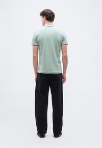 Άνδρας που στέκεται γυρίζοντας την πλάτη φοράει ένα ανοιχτό πράσινο πόλο και μαύρα τζιν, με φόντο ένα απλό ανοιχτόχρωμο φόντο.