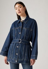 Jachetă din denim cu talie subțire, mâneci lungi și două buzunare la piept. Spalare albastră închisă cu un design structurat și detalii din metal argintiu.