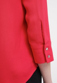 Blusa roja de textura suave, con mangas de tres cuartos y detalle de botones en el puño. El dobladillo tiene una ligera curva.