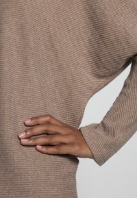 Hand auf der Hüfte ruhend, trägt einen beigen ribbeligen Pullover mit langen Ärmeln, der die Textur zeigt und in einer entspannten Pose vor einem neutralen Hintergrund dargestellt ist.
