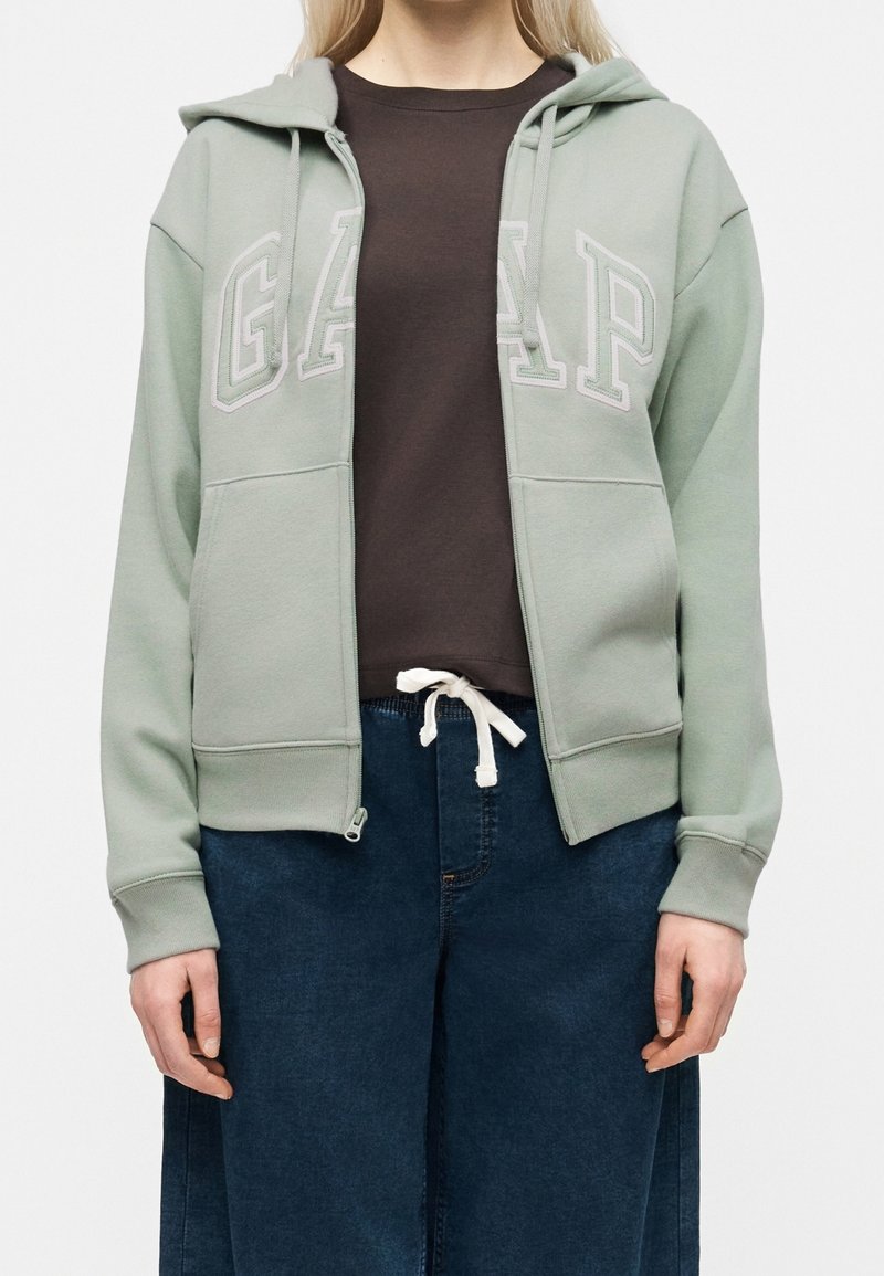 Personne portant un sweat à capuche zippé GAP vert clair sur un t-shirt marron foncé et un pantalon bleu à jambes larges avec une ceinture à cordon blanche.