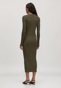 Vrouw met lang krullend haar, gekleed in een aansluitende, lange mouw, geribbelde olijfgroene midi-jurk en zwarte hakken met slingback, bekeken van achteren.