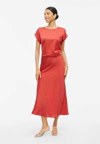 Vestido midi de satén rojo con mangas cortas y enroladas, y un ajuste relajado. Textura suave con un ligero brillo. Combinado con sandalias de tacón transparente.