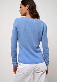 Žena s dlouhými hnědými vlasy, oblečená v světle modrém svetru a bílých kalhotách, otočená zády k jednobarevnému světlému pozadí.