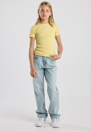 T-shirt giallo a righe con maniche corte e una grafica bianca, abbinato a jeans in denim chiaro e sneakers bianche. Look semplice e rilassato.