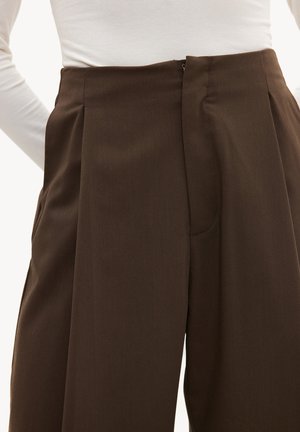 Pantalon marron taille haute et cintré avec plis, porté avec un haut blanc à manches longues rentré, gros plan sur la taille et les hanches.