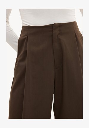 Pantalon marron taille haute et cintré avec plis, porté avec un haut blanc à manches longues rentré, gros plan sur la taille et les hanches.