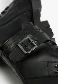Schwarze Lederstiefeletten mit einem seitlichen Reißverschluss, einem Riemen mit einer metallischen Schnalle und dekorativen Nieten auf dem oberen Bereich. Flache Gummisohle.