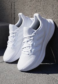 Paire de baskets blanches avec lacets posées sur une surface en béton texturée, une chaussure légèrement appuyée contre un mur.