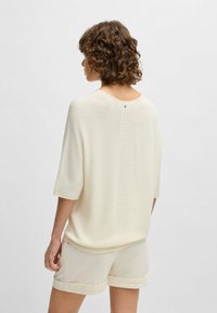 Light cream knitted top with short sleeves, round neckline, and a decorative button detail at the back. Paired with light shorts.