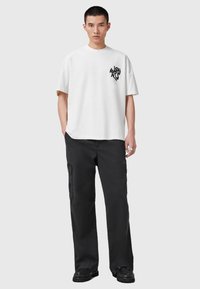 Camiseta blanca de algodón con un gráfico negro en el lado izquierdo del pecho, combinada con pantalones cargo negros de corte holgado que cuentan con bolsillos laterales.