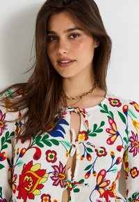 Blusa con estampado floral que presenta diseños coloridos en rojo, amarillo y azul sobre un fondo blanco, con cierre de lazo y un cuello redondo.
