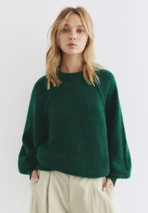 Pull en laine vert avec une coupe ample, des manches raglan et une surface texturée. Associé à un pantalon beige clair.