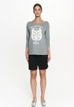 Szary sweatshirt z grafiką białego wilka, z rękawami o długości trzech czwartych, zestawiony z czarnymi szortami. Modelka nosi srebrne baletki.