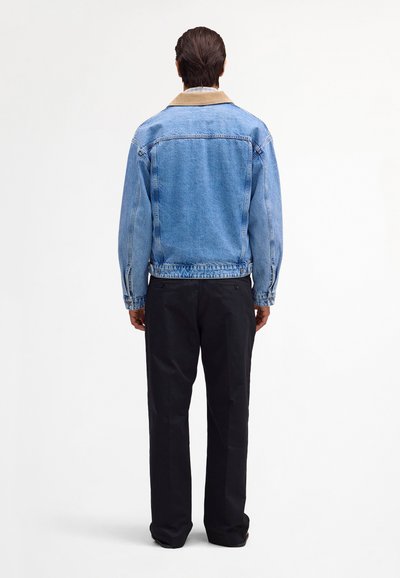 Ein Mann mit dem Rücken zur Kamera trägt eine hellblaue Jeansjacke mit hellbraunem Kragen über einer schwarzen Hose und dunklen Schuhen, steht vor einem schlichten weißen Hintergrund.