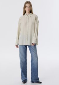 Lichtbeige overhemd met knopen en lange mouwen, met een gestructureerde afwerking, gecombineerd met een blauwe broek met wijde pijpen en verspreide witte spikkels.