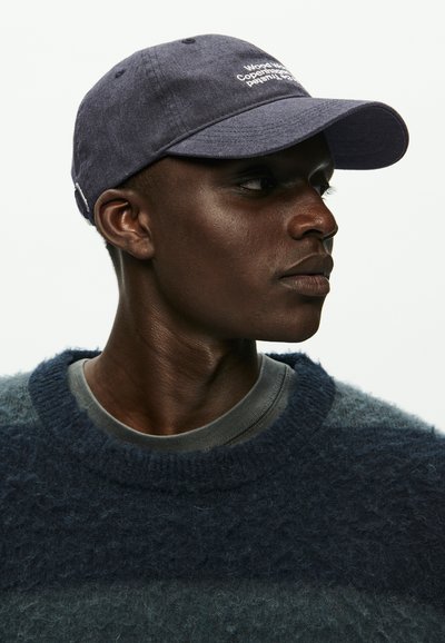 Jūros pajėgų kepurė su siuvinėtu tekstu, pagaminta iš minkštos medžiagos. Turi kreivą šilko kraštą ir reguliuojamą dirželį. Modelis vilki purų jūros pajėgų megztinį.