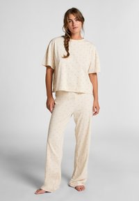 Woman with braided hair wearing cream-colored loose pajama set with small palm tree print, standing barefoot against light gray background.
