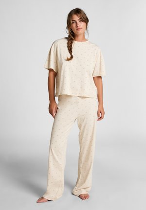 Woman with braided hair wearing cream-colored loose pajama set with small palm tree print, standing barefoot against light gray background.