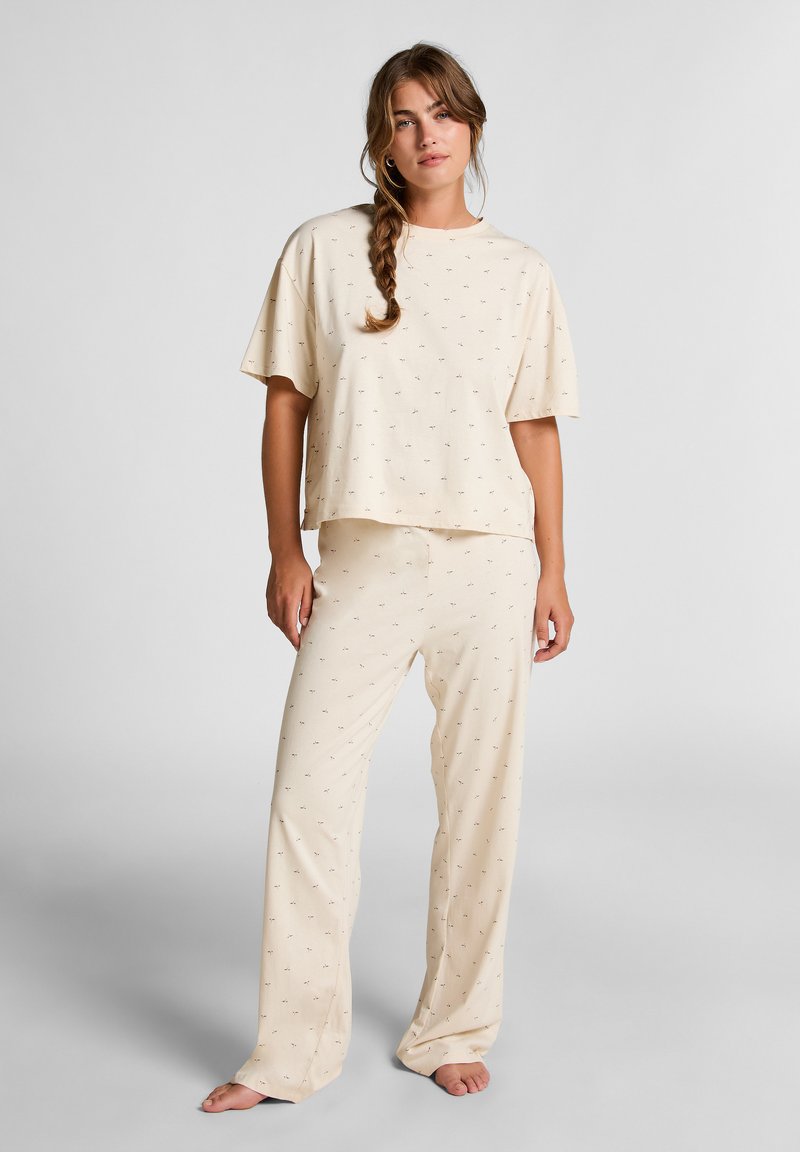 Woman with braided hair wearing cream-colored loose pajama set with small palm tree print, standing barefoot against light gray background.