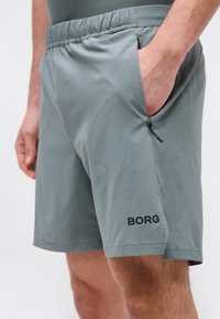Άνδρας που φοράει ανοιχτό γκρι αθλητική βερμούδα με ελαστική μέση και πλαϊνή τσέπη με φερμουάρ, με την ένδειξη «BORG» στο αριστερό πόδι.