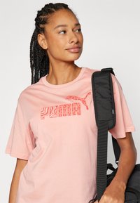 Šviesiai rožinė medvilninė marškinė su ryškiu raudonu "PUMA" logotipu, turinčiu tekstūruotą raštą. Trumpi rankovės ir laisvas kirpimas. Juoda reguliuojama petneša.