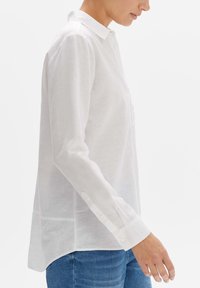 Camicia bianca a maniche lunghe in tessuto leggero con colletto, polsini abbottonati e orlo curvo, abbinata a jeans blu.