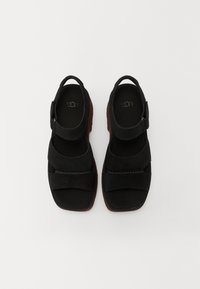 Een paar zwarte suede sandalen met vierkante neuzen, verstelbare enkelbandjes en bruine rubbere zolen, van bovenaf bekeken op een witte achtergrond.