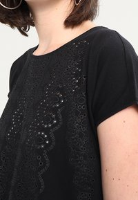 Musta lyhythihainen paita, jossa on monimutkaista reikäkoristetta edessä, pehmeää kangasta ja pyöreä pääntie.