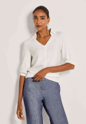 Vrouw die een witte V-hals blouse met driekwartmouwen en een blauwe high-waist spijkerbroek draagt, poserend tegen een effen lichte achtergrond.