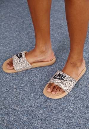 Voeten met beige Nike-slippers met zwart logo op een blauw tapijt met structuur.