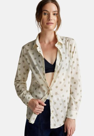 Chemise boutonnée de couleur claire avec un motif floral en vert et rose, munie d'un col et de manches longues, portée par-dessus un top noir.