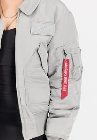 Giubbotto bomber grigio chiaro con texture trapuntata, dotato di un emblema frontale, numerose tasche e un'etichetta rossa "Rimuovere Prima del Volo".