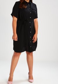 Czarna sukienka z krótkimi rękawami i guzikami, z paskiem w talii i dwiema kieszeniami na piersi, wykonana z lekkiego materiału, zestawiona z różowymi szpilkami.