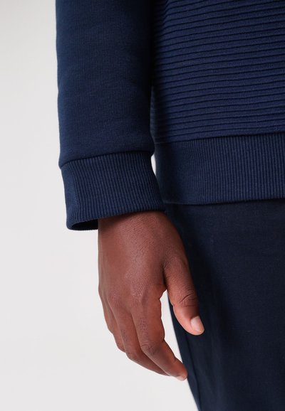 Tumši zila džemperi detaļas ar ribotiem aprocēm un horizontālām teksturētām svītrām uz ķermeņa. Roka daļēji redzama pret neitrālu fonu.