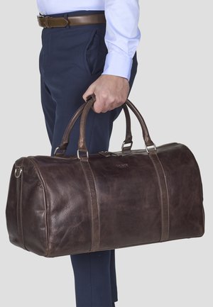 Dark brown leather duffel bag with two handles, silver hardware, and contrasting lighter leather stripes, held by a person in blue pants.