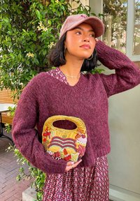 Un pull bordeaux en peluche, une robe à motifs floraux et un sac à main en velours jaune avec des accents de perles. Un feuillage vert en arrière-plan.
