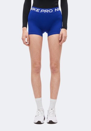 Personne portant un short Nike Pro bleu, des chaussettes blanches, des baskets Nike blanches et un haut à manches longues noir, debout devant un fond uni.