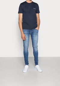 T-shirt in cotone navy con colletto a giro e piccolo logo bianco, abbinato a jeans skinny azzurri e sneakers bianche. Design semplice e aderente.