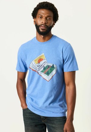 Mann mit Bart, der ein blaues T-Shirt mit einer Grafik eines offenen Buches trägt, das Berge, Bäume und einen Radfahrer auf einem Pfad zeigt.