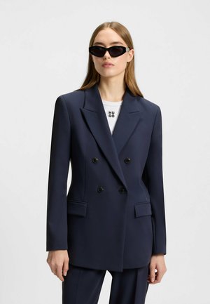 Marineblå dobbeltknap blazer med sorte knapper, slagkraver og sidelommer, parret med en hvid T-shirt med "HUGO" logo. Sorte solbriller.
