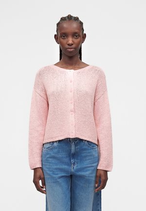 Femme aux cheveux tressés porte un cardigan en maille rose clair boutonné et un jean bleu taille haute sur fond blanc uni.