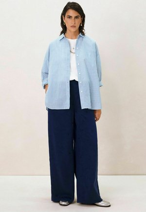 Chemise à rayures bleues surdimensionnée avec des manches retroussées, associée à un pantalon large navy et des chaussures métalliques. Design simple avec une coupe décontractée.