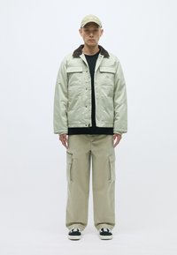 Svetlozelená prešívaná bunda s hnedým zamatovým golierom, čierna košeľa pod ňou a svetlo béžové cargo nohavice. Čierno-biele tenisky.