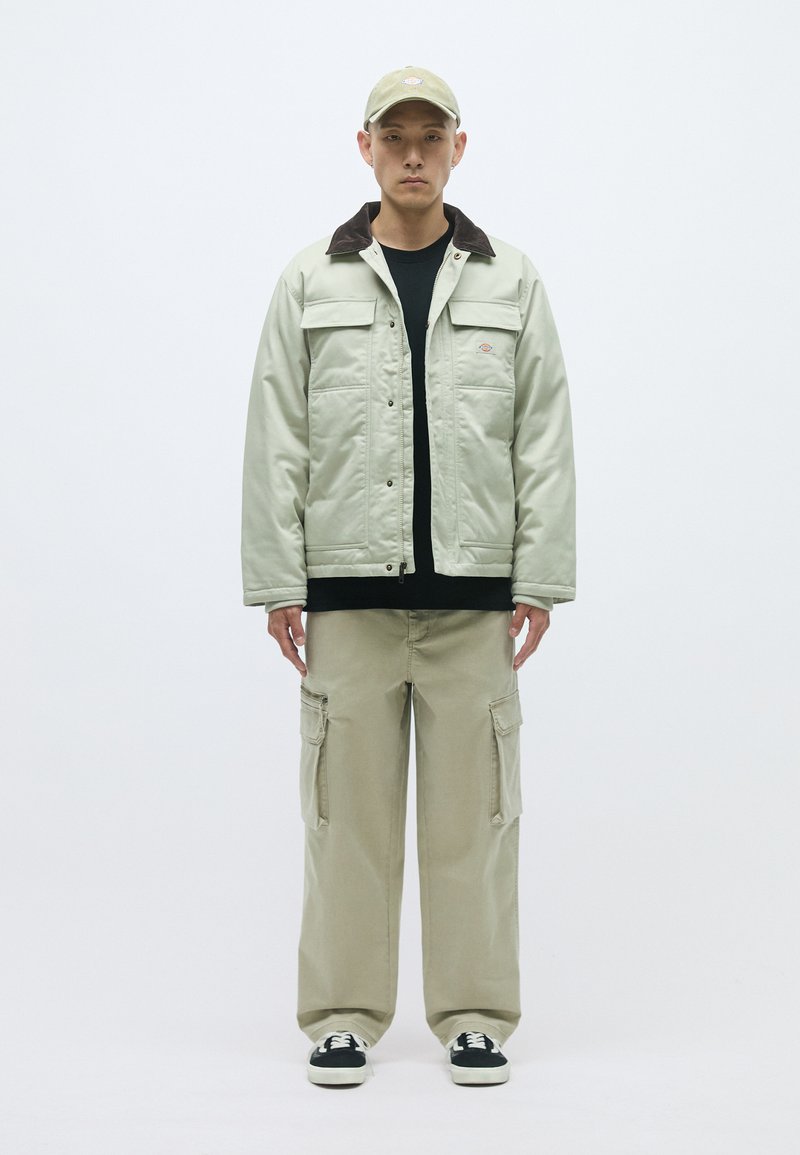 Hellgrüner gesteppter Mantel mit braunem Cordkragen, schwarzes Hemd darunter und hellbeige Cargo-Hosen. Schwarz-weiße Sneaker.